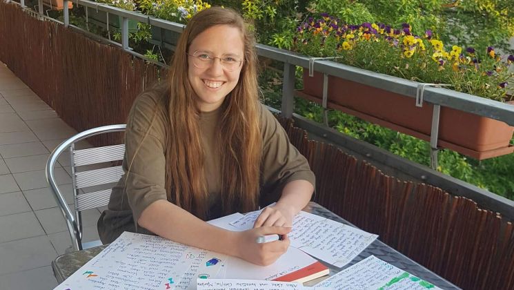 Eine junge Frau mit langen Haaren lächelt, während sie auf einem Balkon mit bunten Blumen und Notizen schreibt.