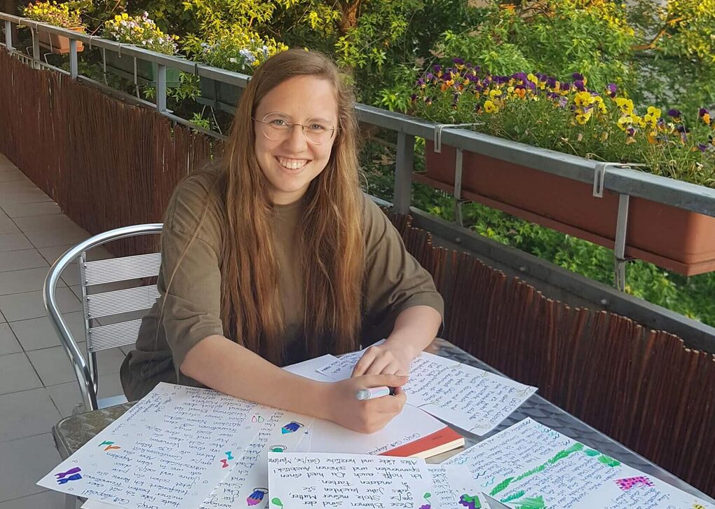 Eine junge Frau mit langen Haaren lächelt, während sie auf einem Balkon mit bunten Blumen und Notizen schreibt.