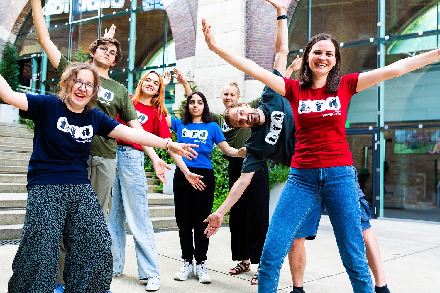 Eine fröhliche Gruppe junger Menschen posiert mit Glück und Ausstrahlung in bunten T-Shirts vor einem architektonischen Hintergrund.