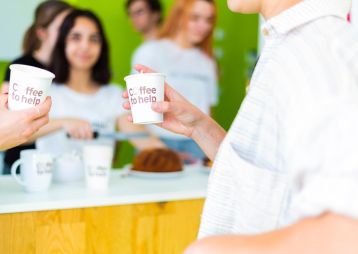 Zwei Personen halten Pappbecher mit 'Coffee to help' vor einer grünen Wand, während im Hintergrund jemand Kaffee zubereitet.