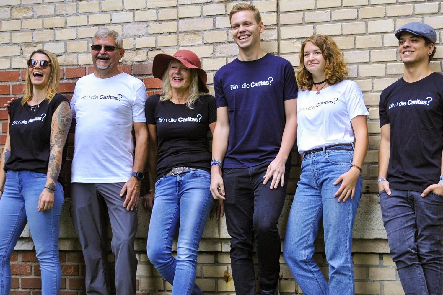 Sechs Personen stehen in einer Reihe, tragen Caritas-T-Shirts und lachen vor einer Backsteinmauer.