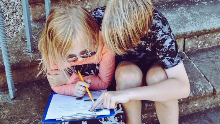 Zwei Kinder sitzen auf Treppenstufen und studieren gemeinsam ein Blatt Papier. Eines von ihnen hält einen Bleistift und zeigt auf etwas.