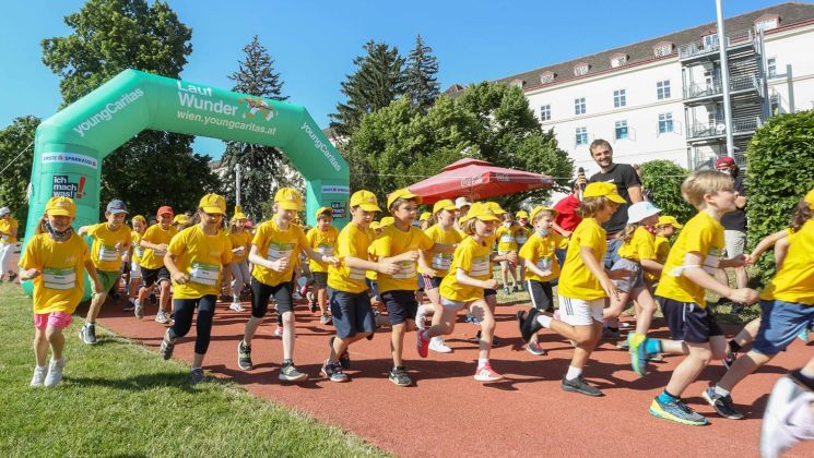 Eine Gruppe von Kindern in gelben T-Shirts und Mützen läuft unter einem grünen Startbogen bei der Veranstaltung Lauf Wunder.