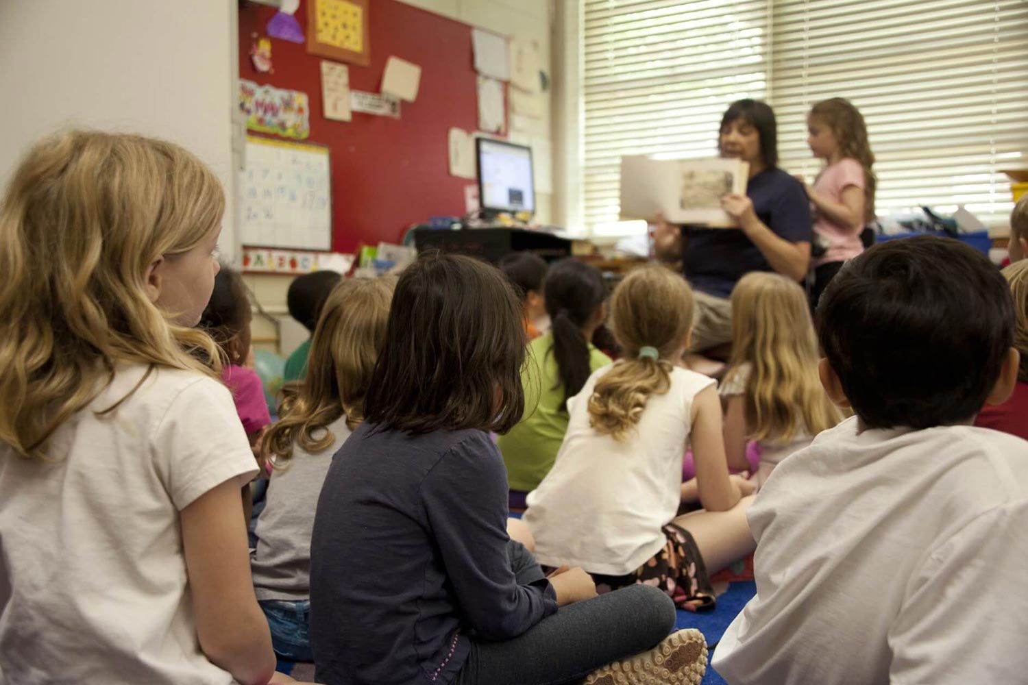 Eine Gruppe von Kindern sitzt im Kreis und hört einer Geschichte zu, die von einer Lehrerin vorgelesen wird.