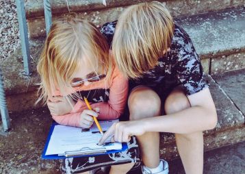 Zwei Kinder sitzen auf Treppenstufen und studieren gemeinsam ein Blatt Papier. Eines von ihnen hält einen Bleistift und zeigt auf etwas.