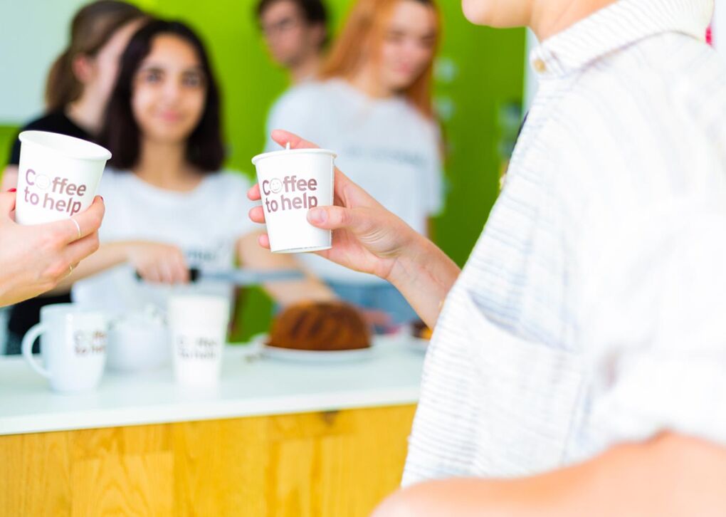 Zwei Personen halten Pappbecher mit 'Coffee to help' vor einer grünen Wand, während im Hintergrund jemand Kaffee zubereitet.