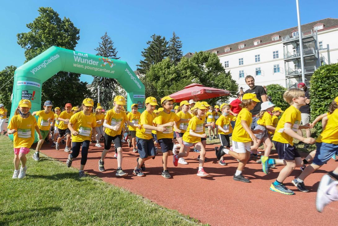 Eine Gruppe von Kindern in gelben T-Shirts und Mützen läuft unter einem grünen Startbogen bei der Veranstaltung Lauf Wunder.