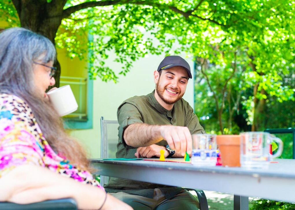 Ein junger Mann lächelt, während er mit bunten Spielsteinen spielt. Eine Frau trinkt aus einer Tasse und sieht ihm zu, umgeben von grünen Bäumen.