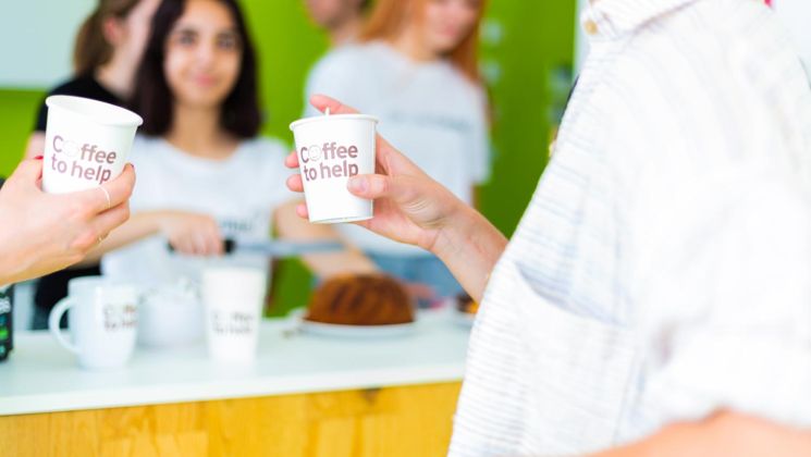 Zwei Personen halten Pappbecher mit 'Coffee to help' vor einer grünen Wand, während im Hintergrund jemand Kaffee zubereitet.