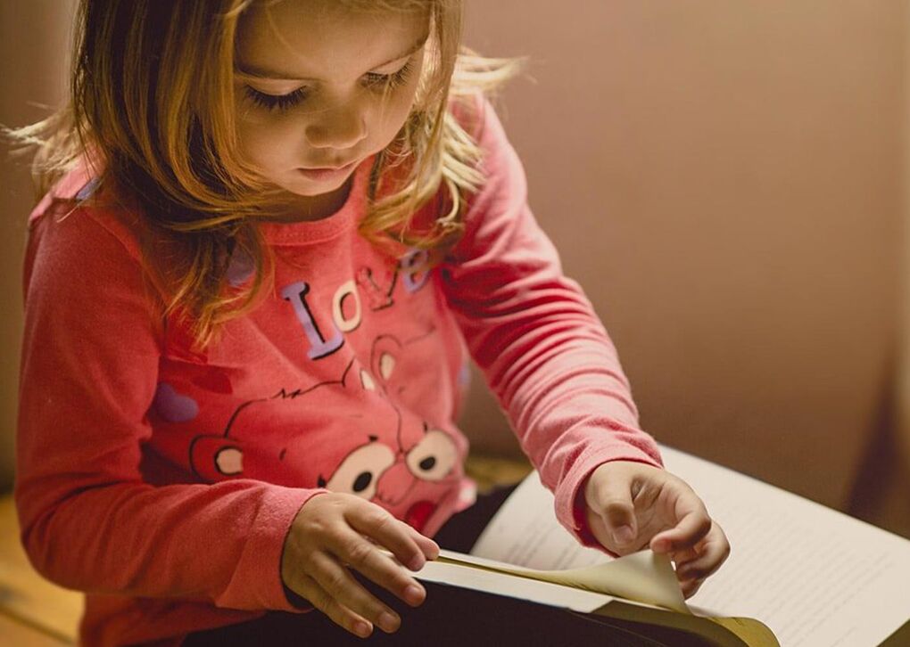 Ein kleines Mädchen in einem rosa Shirt blättert in einem Buch und zeigt Konzentration beim Lesen.