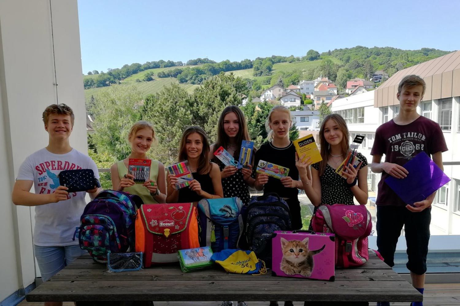 Eine Gruppe von Schülern zeigt stolz ihre Schulmaterialien auf einem Balkon mit grünen Hügeln im Hintergrund.
