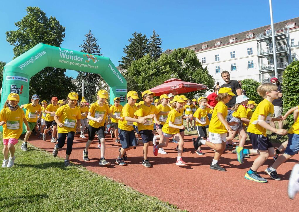 Eine Gruppe von Kindern in gelben T-Shirts und Mützen läuft unter einem grünen Startbogen bei der Veranstaltung LaufWunder.