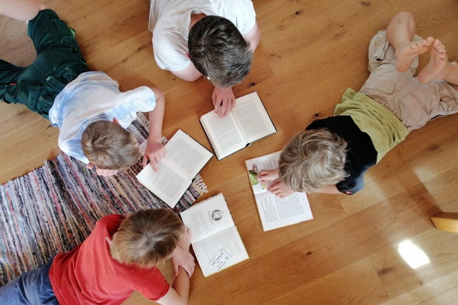 Fünf Kinder liegen auf dem Boden und lesen Bücher. Sie sind um einen bunten Teppich herum aufgestellt.