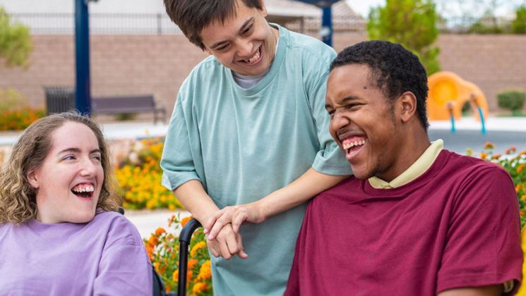 Drei junge Menschen lachen zusammen im Park, umgeben von bunten Blumen. Eine Person steht und hält die Hand eines Freundes. Ein fröhlicher Moment.