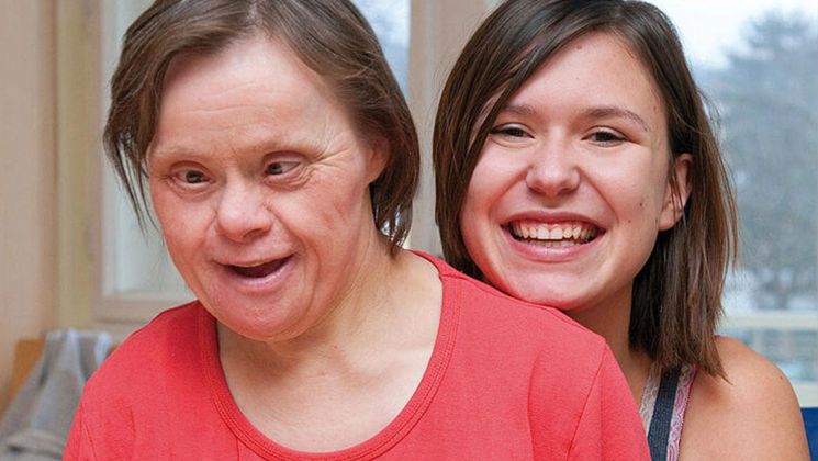 Zwei Frauen lächeln zusammen, eine sitzt vorne mit Down-Syndrom und die andere steht lächelnd dahinter.